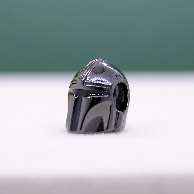 Puede incluir: Un charm met&aacute;lico gris oscuro con forma de casco. El casco tiene una superficie lisa y reflectante con detalles visibles, incluyendo una visera y elementos laterales. El charm est&aacute; dise&ntilde;ado con un agujero para ensartar.