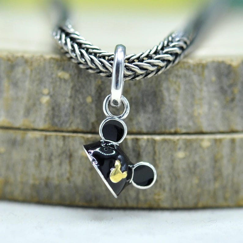 May include: A silver charm with a black and gold Mickey Mouse design. The charm is suspended from a silver chain. The Mickey Mouse charm has black ears and a black hat with a gold emblem.