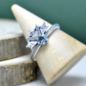 Puede incluir: Un anillo plateado con una piedra central azul claro, rodeada de piedras más pequeñas, transparentes y azul claro. La banda está adornada con pequeñas piedras transparentes. El anillo se exhibe en un soporte de madera.