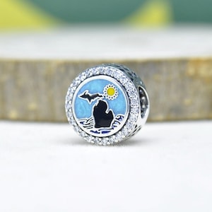 May include: A round silver charm with a blue enamel design featuring a black silhouette of a landmass, a yellow sun, and a flying bird. The charm is surrounded by a ring of clear, sparkling stones.