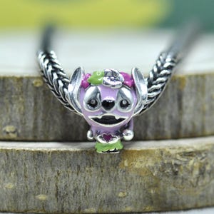 May include: A silver charm bead featuring a stylized purple creature with large ears, a floral crown in pink and green, and a small heart detail on its chest. The charm is threaded onto a silver chain with a braided, textured metal link design.