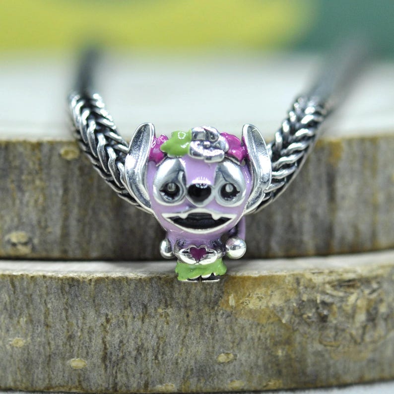 May include: A silver charm bead featuring a stylized purple creature with large ears, a floral crown in pink and green, and a small heart detail on its chest. The charm is threaded onto a silver chain with a braided, textured metal link design.