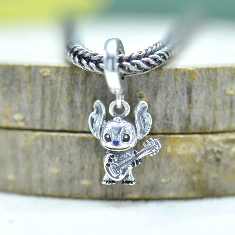 May include: A silver charm bead featuring a detailed figure of the character Stitch from Lilo and Stitch, depicted holding a small guitar. The charm has a polished metallic finish with dark accents on the eyes and nose, suspended from a silver chain.