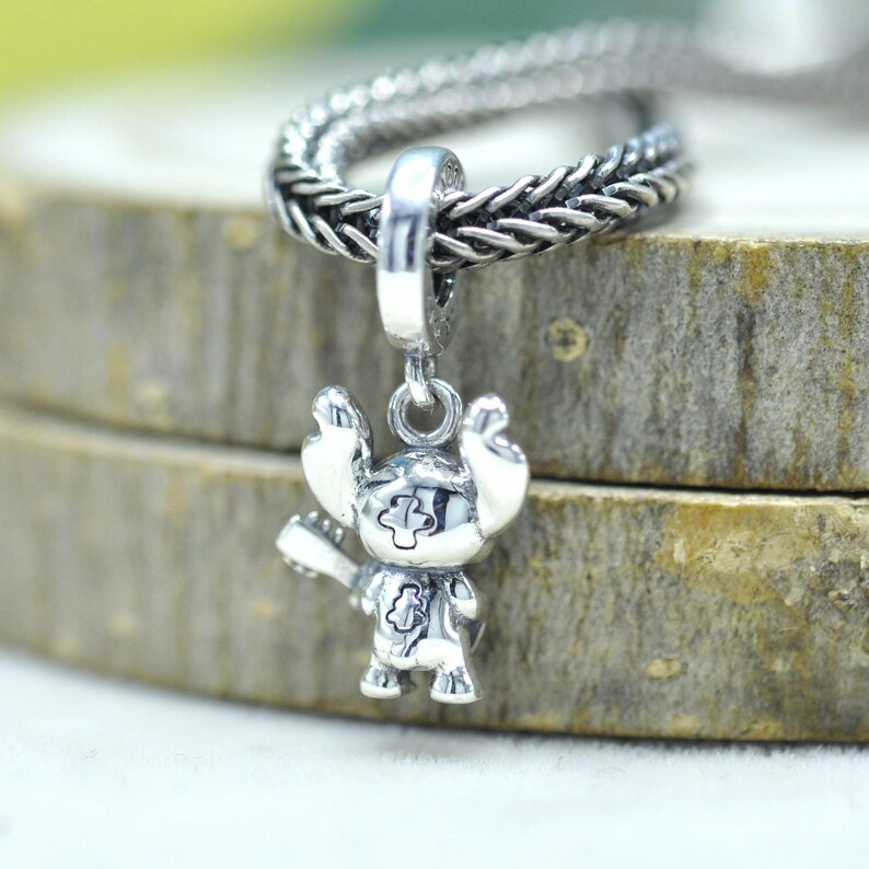 May include: A polished silver charm pendant shaped like a cartoon creature holding a guitar, featuring etched details on the face and body. The charm is attached to a thick, braided silver chain with a metallic finish and a reflective surface.