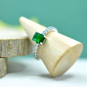 Op de afbeelding: Een zilveren ring met een rechthoekige, smaragdgeslepen groene edelsteen. De ringband is versierd met kleine, heldere edelstenen. De ring wordt gepresenteerd op een lichtgekleurde houten ringhouder.