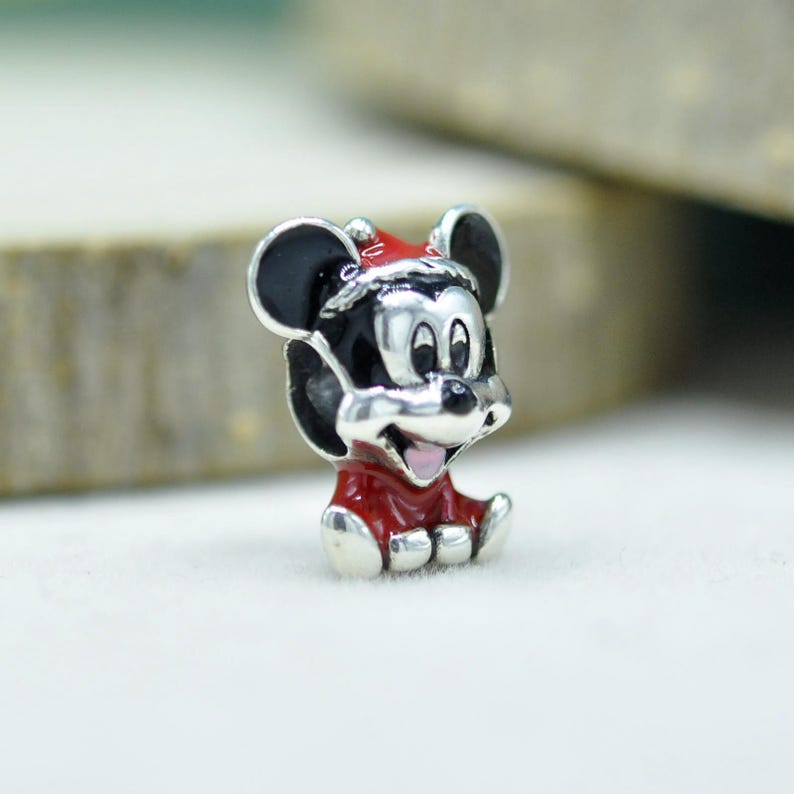 May include: A silver-toned metal charm shaped like a cartoon mouse character wearing a red Santa hat and a red outfit. The charm features black enamel detailing on the ears and head, with a small pink tongue visible in the smiling mouth.