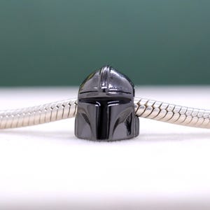Puede incluir: Un charm de color gris oscuro con un dise&ntilde;o de casco mandaloriano. El charm est&aacute; unido a una cadena plateada, mostrando la forma y los detalles distintivos del casco. El casco tiene un acabado brillante y es un art&iacute;culo popular para los fans de la serie.