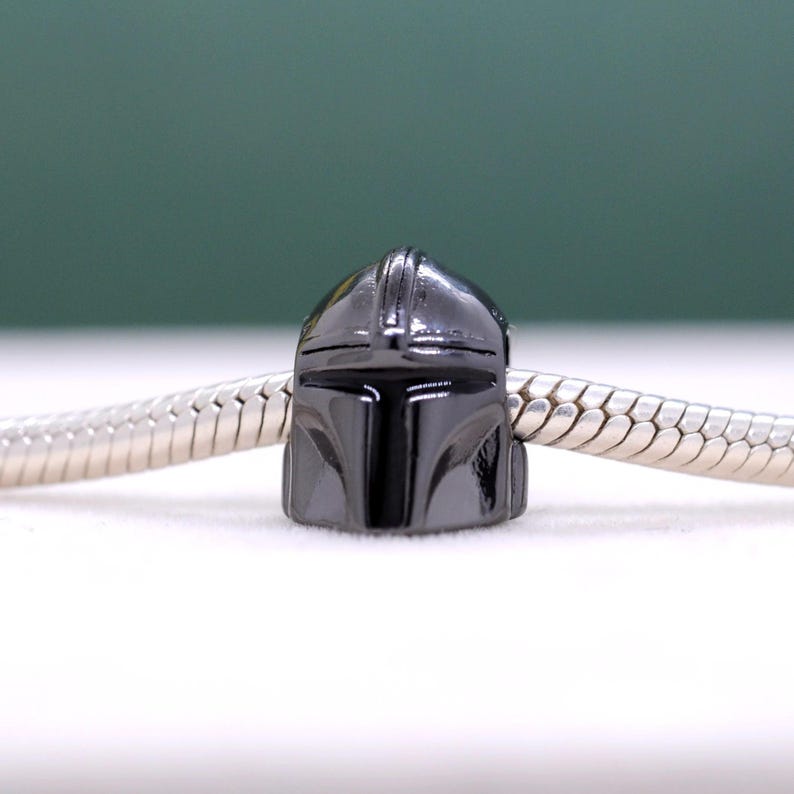 Puede incluir: Un charm de color gris oscuro con un dise&ntilde;o de casco mandaloriano. El charm est&aacute; unido a una cadena plateada, mostrando la forma y los detalles distintivos del casco. El casco tiene un acabado brillante y es un art&iacute;culo popular para los fans de la serie.