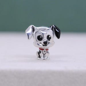 Puede incluir: Un charm de color plateado con forma de perro de dibujos animados. El perro tiene ojos negros, una oreja negra y un collar rojo. El charm está sobre una superficie blanca.