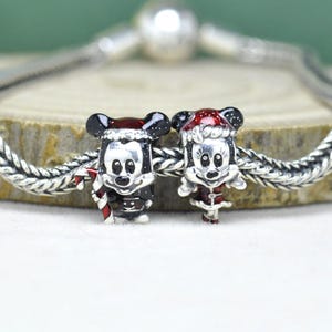 Op de afbeelding: Twee zilveren kerstbedels met Mickey en Minnie Mouse. Mickey draagt een zwarte outfit en een kerstmuts, met een zuurstok. Minnie draagt een rode jurk en een kerstmuts. Beide bedels zitten aan een zilveren ketting.