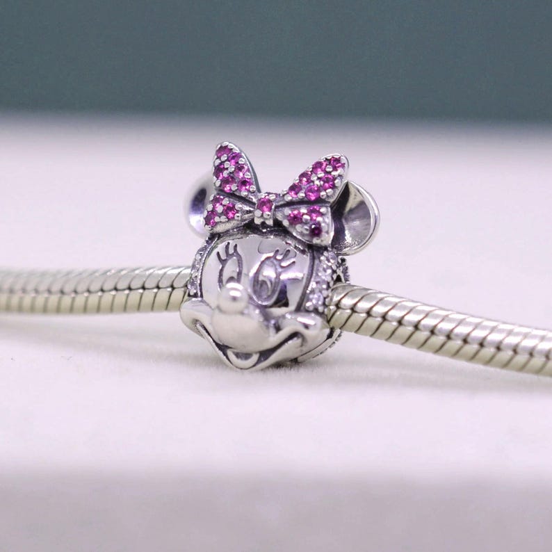 May include: A silver charm featuring Minnie Mouse with a pink bow and sparkling accents. The charm is detailed with a smiling face and is designed to be added to a bracelet or necklace. The charm is shown on a silver chain.