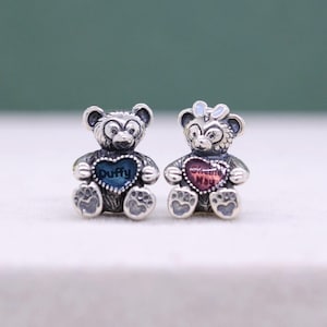 Könnte beinhalten: Zwei silberfarbene Bärenanhänger. Ein Bär hält ein blaues Herz mit dem Wort "Duffy" in Weiß. Der andere Bär hält ein rosa Herz mit Text. Beide Bären haben eine Schleife und pfotenartige Füße. Die Anhänger sind wahrscheinlich für ein Armband oder eine Halskette.