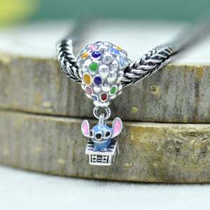 May include: A silver charm bead featuring a cluster of colorful enamel balloons in red, blue, yellow, green, and purple. A small blue character with pink ears sits inside a silver house structure suspended from the balloon cluster on a silver chain.