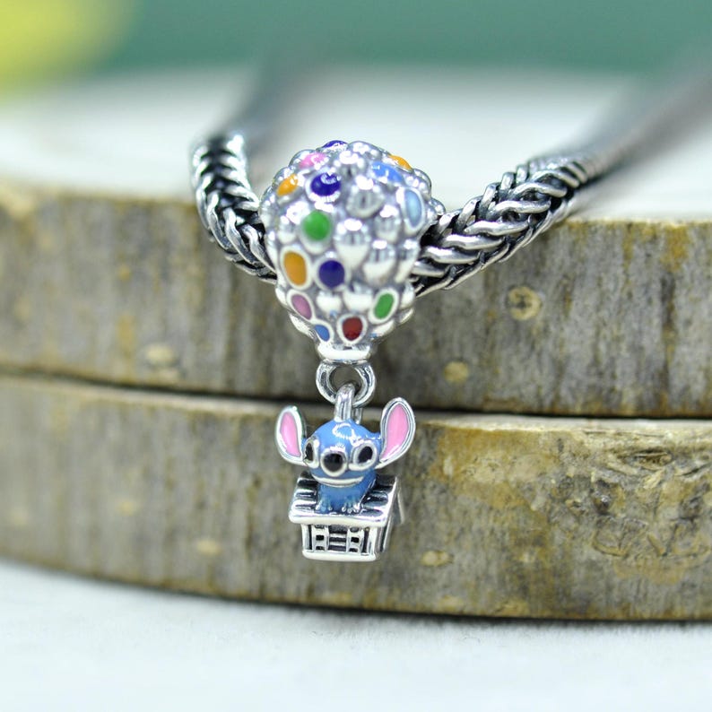 May include: A silver charm bead featuring a cluster of colorful enamel balloons in red, blue, yellow, green, and purple. A small blue character with pink ears sits inside a silver house structure suspended from the balloon cluster on a silver chain.