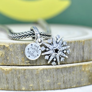Puede incluir: Un charm de copo de nieve plateado con un centro de estrella negra y detalles de cristal transparente. Un pequeño charm redondo con el texto "THE FUTURE IS BRIGHT" cuelga junto al copo de nieve. El charm está unido a una cadena plateada.