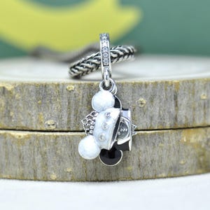 May include: A silver charm with a Mickey Mouse design. The charm features a black and white Mickey Mouse head with a white jacket and black bow tie. The charm is accented with small, clear crystals and hangs from a silver chain.