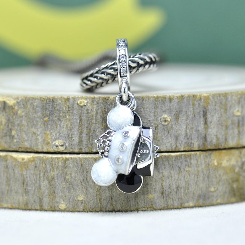 May include: A silver charm with a Mickey Mouse design. The charm features a black and white Mickey Mouse head with a white jacket and black bow tie. The charm is accented with small, clear crystals and hangs from a silver chain.