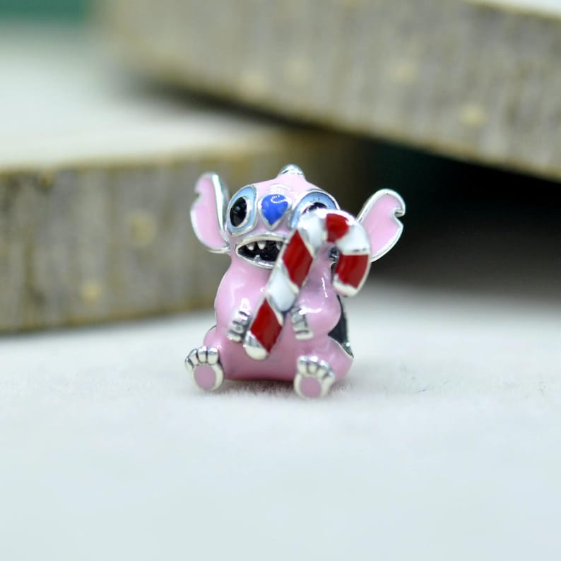 May include: A metal charm bead shaped like a pink creature with large ears, black eyes, and a blue nose, holding a red and white striped candy cane. The charm features enamel detailing and a silver-toned finish, designed for a bracelet or necklace.