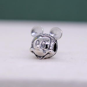 May include: A silver charm featuring the iconic face of Mickey Mouse, complete with round ears and a smiling expression. The charm is detailed with small, sparkling accents around the face, adding a touch of shimmer to the design. This charm is a collectible item.