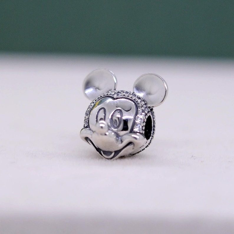 May include: A silver charm featuring the iconic face of Mickey Mouse, complete with round ears and a smiling expression. The charm is detailed with small, sparkling accents around the face, adding a touch of shimmer to the design. This charm is a collectible item.