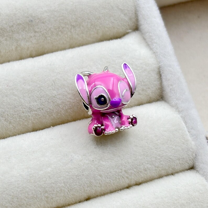May include: A pink and purple enamel charm of a cartoon character with one eye closed, resembling Stitch from the Disney movie Lilo & Stitch.