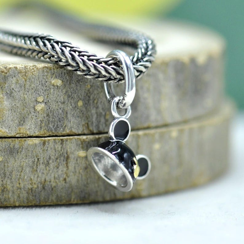 May include: A silver-toned charm with a black and gold teacup design. The charm is attached to a braided silver chain. The teacup has a small handle and a detailed rim. The charm is likely made of sterling silver.