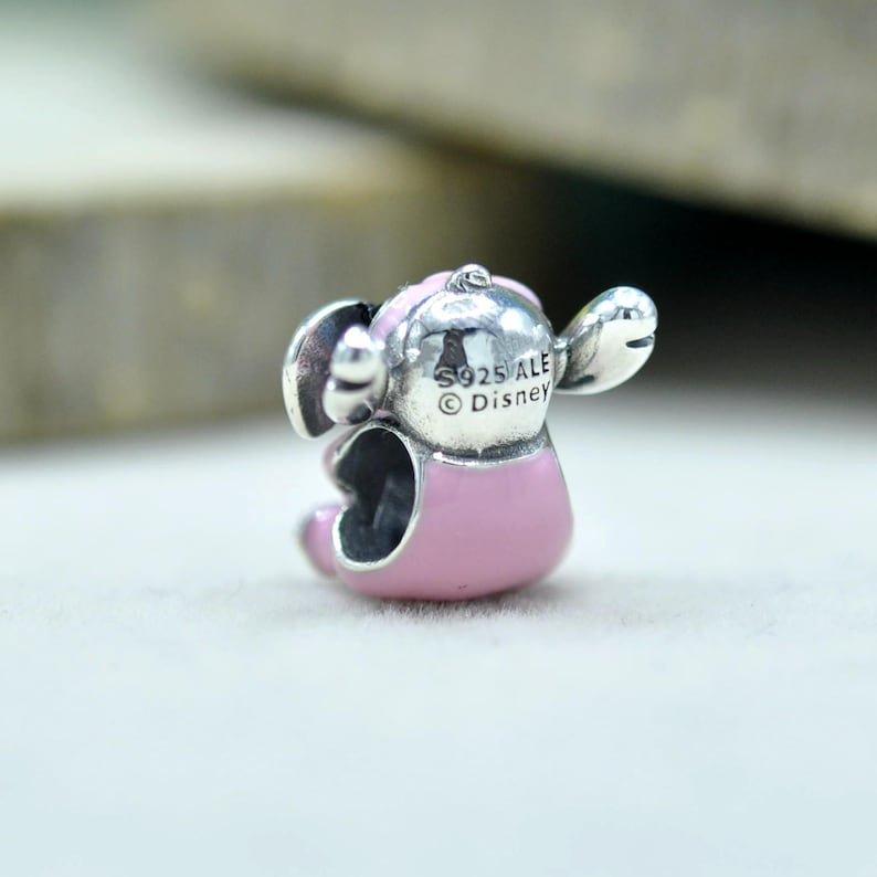 May include: A silver charm bead shaped like a character with large ears and a pink enameled body. The back of the head features engraved text reading S925 ALE and Disney. The charm has a hollow center for threading onto a bracelet or necklace chain.