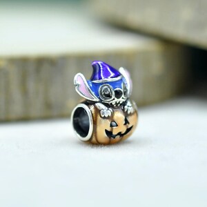 May include: A silver charm bead featuring a blue character with large ears wearing a purple pointed hat, sitting inside a carved orange pumpkin with a black jack-o'-lantern face. The charm has a hollow center for threading onto a bracelet.