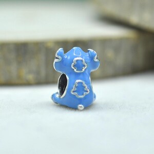 May include: A light blue enamel charm bead featuring a stylized character design with silver-toned outlines. The bead has a central opening for a bracelet and displays a smooth, glossy finish with distinct embossed shapes on its surface.