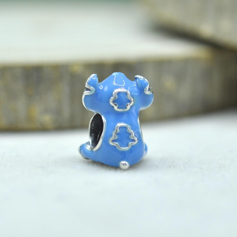 May include: A light blue enamel charm bead featuring a stylized character design with silver-toned outlines. The bead has a central opening for a bracelet and displays a smooth, glossy finish with distinct embossed shapes on its surface.