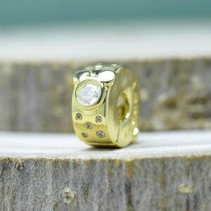 Puede incluir: Un charm dorado con un dise&ntilde;o de cabeza de Mickey Mouse. El charm presenta una gema redonda grande y transparente y varias gemas m&aacute;s peque&ntilde;as y transparentes. El charm est&aacute; sobre una superficie de madera.