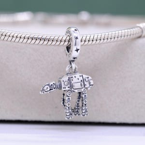 Puede incluir: Un charm de plata que representa un caminante AT-AT de Star Wars. El charm está detallado con características mecánicas y cuelga de una cadena de plata. El charm está diseñado para ser un colgante o una cuenta para una pulsera.