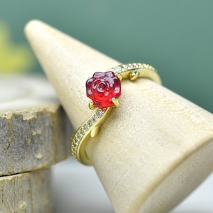 Könnte beinhalten: Ein goldfarbener Ring mit einem roten rosenförmigen Edelstein. Das Band ist mit kleinen, klaren Edelsteinen besetzt und hat ein zartes, rankenartiges Design. Der Ring wird auf einem hellen Holzringhalter präsentiert.
