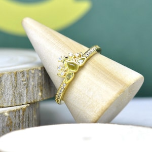 Op de afbeelding: Een goudkleurige ring met een bloemmotief, met een olijfgroene edelsteen en kleine heldere stenen. De ring wordt gepresenteerd op een houten kegelvormige standaard, die de details benadrukt.