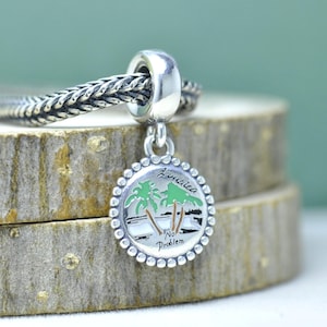 Könnte beinhalten: Ein silberner Charm-Anhänger mit einem runden Anhänger, der eine tropische Szene mit Palmen und den Worten "Jamaica" und "No Problem" zeigt. Der Charm ist an einer Silberkette befestigt und der Anhänger hat einen Perlenrand.