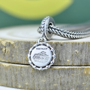 Może przedstawiać: Srebrny charms z szczegółową ilustracją budynku, z napisem "VIETNAM". Okrągły charms jest obramowany akcentami w kształcie serca i wisi na plecionym srebrnym łańcuszku. Charms jest przymocowany do srebrnej bransoletki.