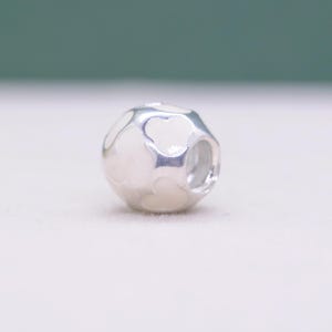 May include: A silver charm with a white enamel design. The charm has a spherical shape with a series of heart-shaped cutouts. The white enamel fills the spaces between the cutouts, creating a contrasting pattern.