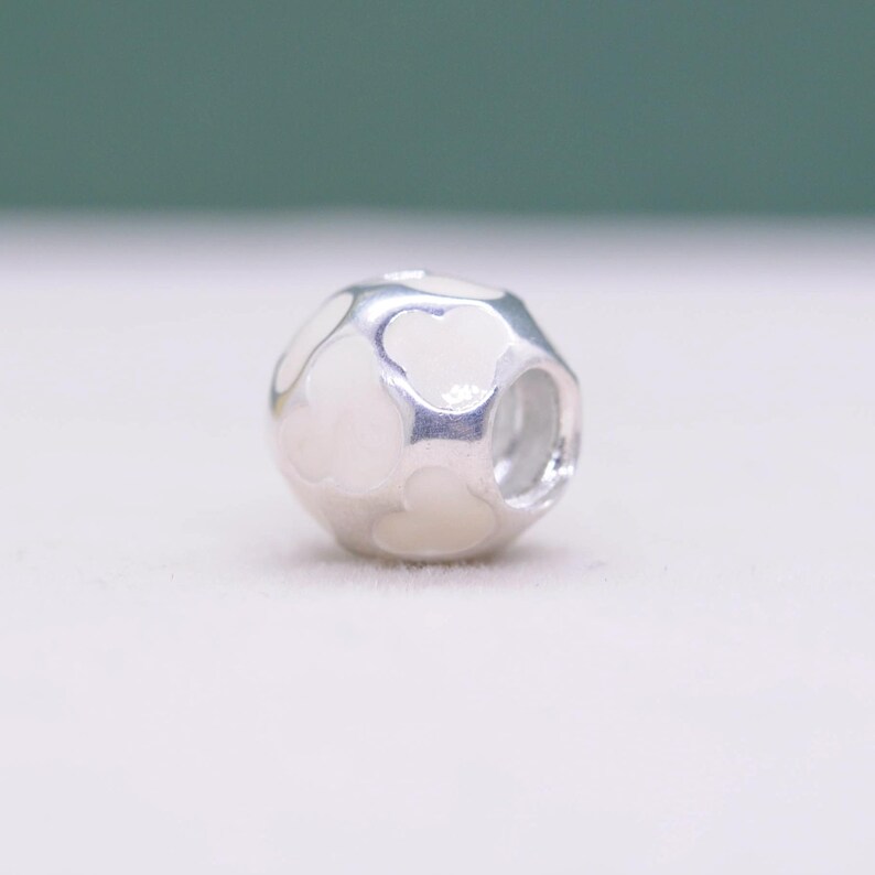 May include: A silver charm with a white enamel design. The charm has a spherical shape with a series of heart-shaped cutouts. The white enamel fills the spaces between the cutouts, creating a contrasting pattern.