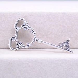 May include: A silver-colored key pendant with a castle design at the end. The key's head features a Mickey Mouse-inspired design with clear gemstones. The key's handle is a straight bar with a detailed castle design.