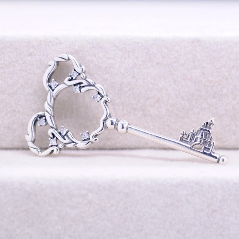 May include: A silver-colored key pendant with a castle design at the end. The key's head features a Mickey Mouse-inspired design with clear gemstones. The key's handle is a straight bar with a detailed castle design.