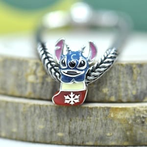 Puede incluir: Un dije de metal plateado con un personaje azul de orejas grandes e interiores rosas, que emerge de un calcet&iacute;n navide&ntilde;o crema y rojo decorado con un copo de nieve blanco, unido a una banda de metal plateado con dise&ntilde;o trenzado.
