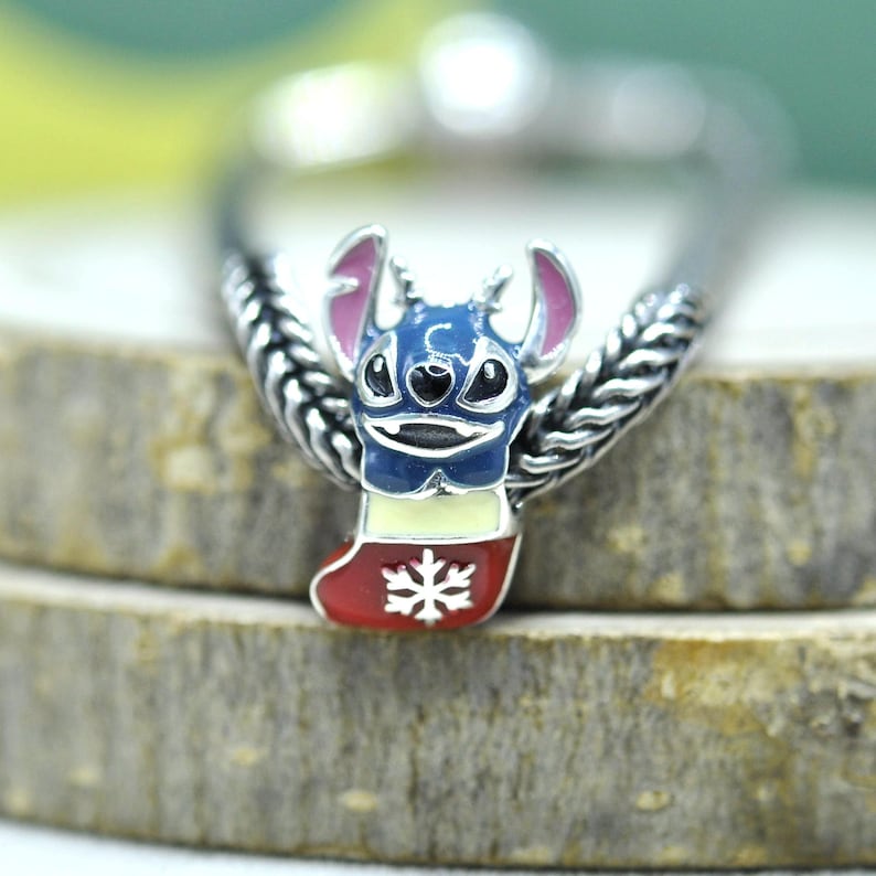 Puede incluir: Un dije de metal plateado con un personaje azul de orejas grandes e interiores rosas, que emerge de un calcet&iacute;n navide&ntilde;o crema y rojo decorado con un copo de nieve blanco, unido a una banda de metal plateado con dise&ntilde;o trenzado.