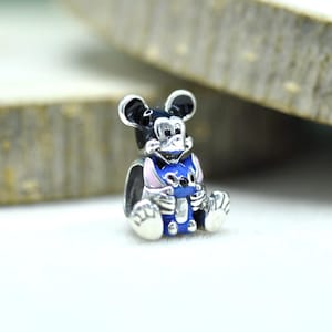 Puede incluir: Un abalorio de plata que muestra una figura de Mickey Mouse en blanco y negro sosteniendo a un peque&ntilde;o personaje de Stitch en azul y rosa. El dije de metal tiene un acabado pulido con detalles en esmalte y est&aacute; dise&ntilde;ado para una pulsera.