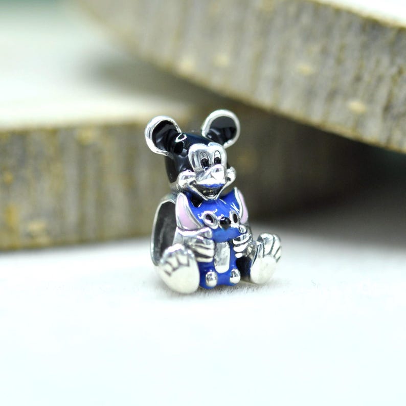 Puede incluir: Un abalorio de plata que muestra una figura de Mickey Mouse en blanco y negro sosteniendo a un peque&ntilde;o personaje de Stitch en azul y rosa. El dije de metal tiene un acabado pulido con detalles en esmalte y est&aacute; dise&ntilde;ado para una pulsera.
