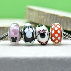 以下が含まれることがあります： シルバーのアクセントが付いた4つのガラスビーズ。ビーズはシルバーのチェーンに付いています。最初のビーズはピンク色で、ミッキーマウスのデザインです。2番目のビーズは白と黒で、ミッキーマウスのデザインです。3番目のビーズは白で、ミッキーマウスのデザインです。4番目のビーズはオレンジ色で、白い水玉模様です。