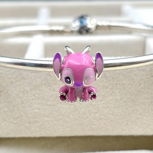 May include: A pink and purple enamel charm of Stitch, a character from the Disney movie Lilo & Stitch, with a winking eye. The charm is made of silver and has a detailed design.