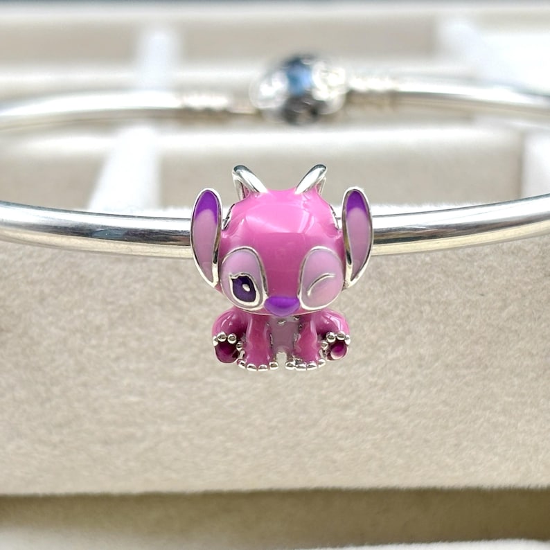 May include: A pink and purple enamel charm of Stitch, a character from the Disney movie Lilo & Stitch, with a winking eye. The charm is made of silver and has a detailed design.