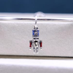 May include: Silver charm shaped like a nutcracker soldier. The charm is wearing a blue and white hat with a star on top. The soldier is holding a red and white weapon in each hand.