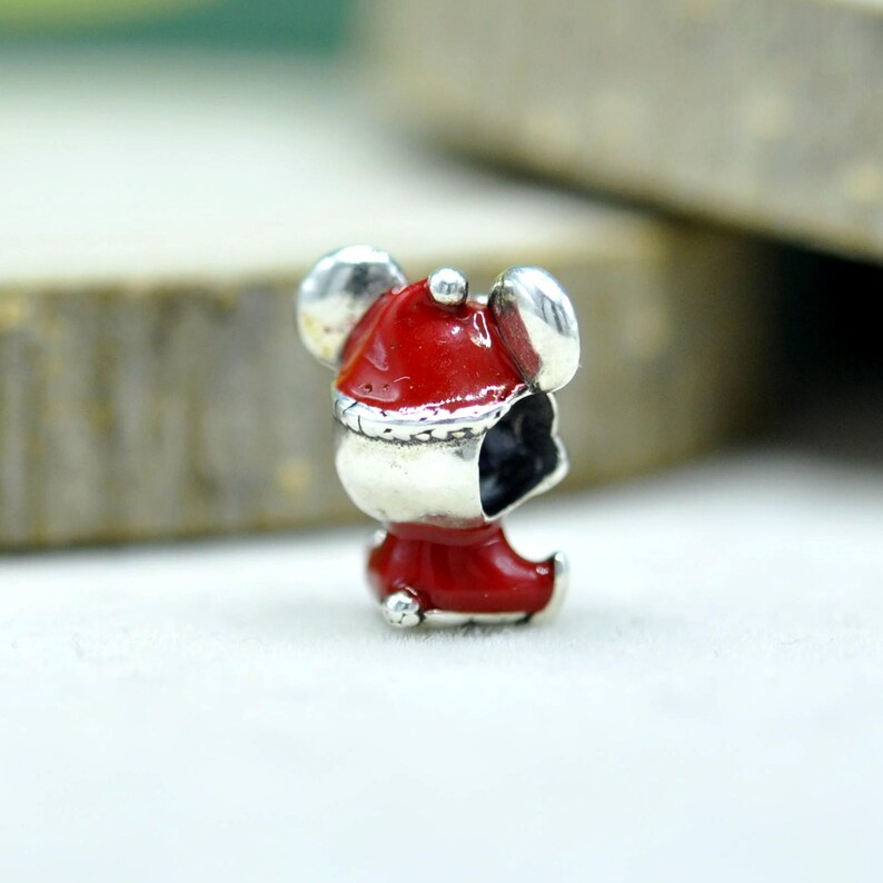 May include: A silver charm bead shaped like a mouse wearing a red enamel Santa hat and a red enamel scarf. The mouse has polished silver ears and a small silver tail, with a central hole for threading onto a bracelet or necklace chain.