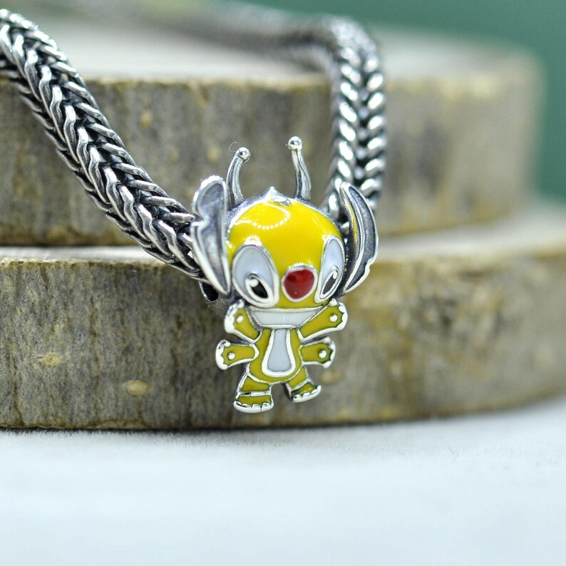 May include: A silver charm bead featuring a yellow and white enameled alien character with large ears, antennae, and a red nose, threaded onto a silver chain with a woven texture. The character has four arms and is depicted in a standing position.
